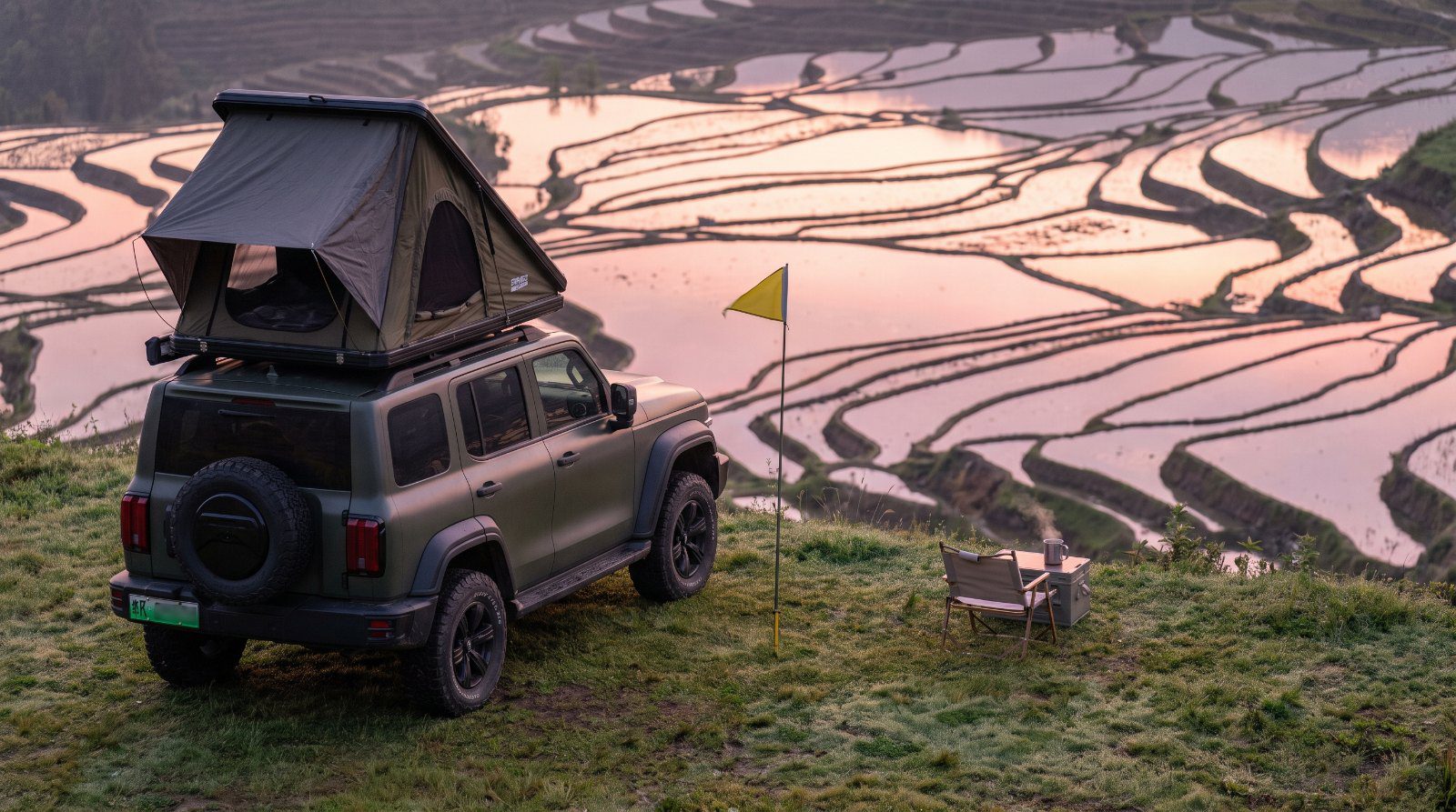 Fuoristrada con tenda sul tetto davanti alle risaie a terrazza dello Yunnan, con una bandierina gialla da guida turistica piantata nel prato